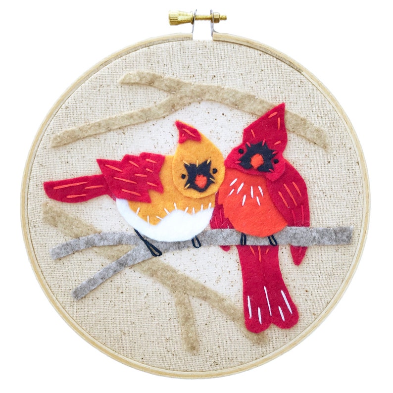 May include: A felt embroidery hoop with two red cardinals perched on a branch. The cardinals are stitched with white and orange details. The background is a beige fabric with a subtle texture.