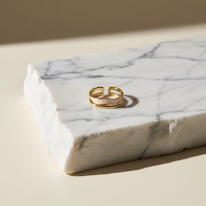 May include: A gold ring with a light pink enamel inlay. The ring features a double-band design and a smooth, polished finish. The ring is displayed on a white marble surface.