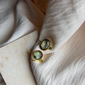 May include: A pair of gold-toned earrings featuring a large, faceted, pale green oval gemstone paired with a smaller, round, vibrant green gemstone. The earrings have a curved wire hook design and a polished metal setting surrounding the stones.