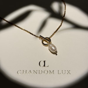 Puede incluir: Un collar dorado con cierre de palanca y una perla blanca colgante. El collar está sobre una superficie blanca con el texto dorado "CHANDOM LUX". La cadena tiene un diseño delicado.