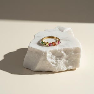 May include: A gold ring with a row of colorful, rectangular gemstones. The gemstones include green, pink, orange, and clear stones. The ring is displayed on a white, textured stone against a neutral background.