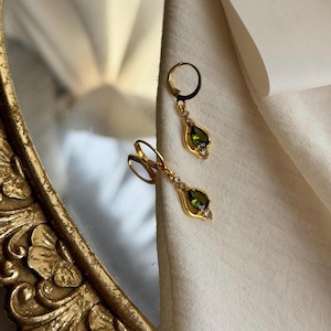 May include: A pair of gold hoop earrings featuring a dangling teardrop-shaped olive green gemstone. Each earring is accented with a small clear stone at the top and bottom of the teardrop setting, creating a delicate and refined jewellery piece.