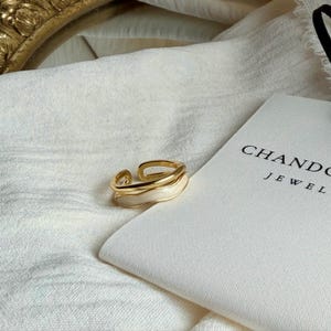 May include: A gold-toned ring with a white enamel inlay. The ring has a layered design and is set against a white background. The text "CHANDO JEWEL" is visible on a white pouch.