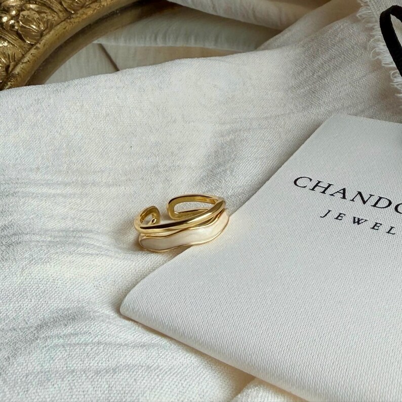 May include: A gold-toned ring with a white enamel inlay. The ring has a layered design and is set against a white background. The text "CHANDO JEWEL" is visible on a white pouch.