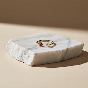 May include: A gold open ring featuring a red gemstone and a white gemstone. The ring is presented on a white marble block, set against a neutral backdrop. Jewellery.