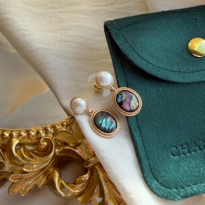 Op de afbeelding: Een paar oorbellen met een ronde witte parel bevestigd aan een ovaalvormige hanger met een donkere, iriserende abalone schelp inlay. De hanger is omlijst door een getextureerde goudkleurige metalen rand en de oorbellen zijn voorzien van doorzichtige sluitingen.