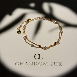 Può includere: Un delicato braccialetto dorato a due fili, ornato da piccole perle e perline scure. Il braccialetto è esposto su una superficie bianca con il nome del marchio "CHANDOM LUX" stampato in oro.