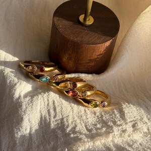Op de afbeelding: Een verzameling goudkleurige ringen, elk met een gekleurde edelsteen. De edelstenen zijn helder, rood, blauw, roze, geel en groen. De ringen worden gepresenteerd op een wit oppervlak met een houten standaard op de achtergrond.