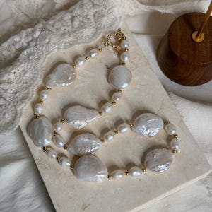 Op de afbeelding: Een ketting met grote, onregelmatige witte barokparels afgewisseld met kleine, ronde goudkleurige kraaltjes. Het sieraad is voorzien van een ronde knevelsluiting van goudkleurig metaal, wat een doorlopende cirkel van glanzende witte parels vormt.