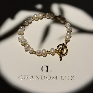 Pode incluir: Uma pulseira com pequenas pérolas de água doce brancas e irregulares intercaladas com minúsculas contas douradas. A peça é fixada com um fecho de barra e anel dourado. O nome da marca CHANDOM LUX e um logótipo CL estilizado estão impressos.