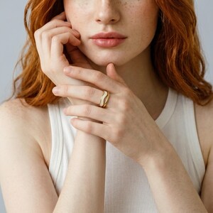 May include: A gold-toned ring with a white enamel wave design. The ring is a simple band with a modern, minimalist aesthetic. The white enamel contrasts with the gold, creating a striking visual effect. The ring is a piece of jewelry.