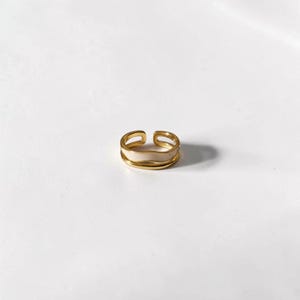 May include: A gold-colored adjustable ring with a cream-colored enamel band. The ring has a double-band design and is set against a white background. This piece of jewelry is suitable for everyday wear.