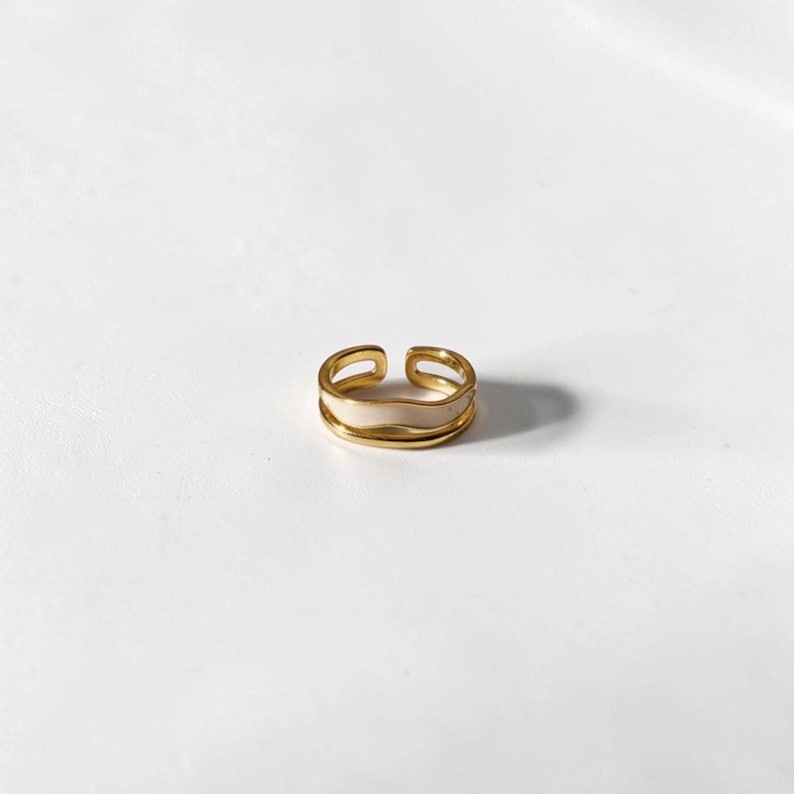 May include: A gold-colored adjustable ring with a cream-colored enamel band. The ring has a double-band design and is set against a white background. This piece of jewelry is suitable for everyday wear.