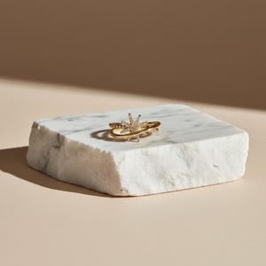 Op de afbeelding: Een goudkleurige ring met een sterrenpatroon, bezet met kleine, heldere edelstenen. De ring ligt op een witte marmeren blok, wat de ingewikkelde details benadrukt. Het ontwerp van de ring omvat een band en een sterrenpatroon.