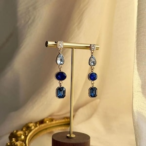May include: A pair of gold-tone drop earrings featuring a vertical arrangement of three distinct blue and clear crystals. Each earring consists of a top clear rectangular stone, a middle clear teardrop, a dark blue oval, and a bottom dark blue rectangle.