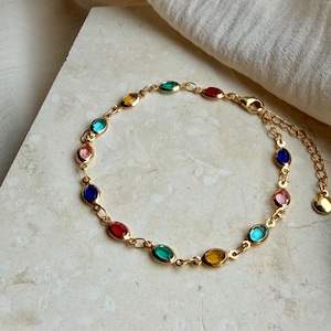 Puede incluir: Una pulsera de cadena en tono dorado que presenta una serie de pequeñas piedras facetadas de forma ovalada y multicolor. Las piedras están engastadas en marcos metálicos dorados individuales e incluyen tonos rojos, azules, verdes, amarillos y rosas.
