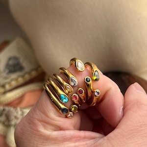 Könnte beinhalten: Eine Sammlung goldfarbener Ringe, jeder mit einem einzigartigen Edelstein in verschiedenen Farben. Zu den Edelsteinen gehören klar, blau, rot, grün und rosa. Die Ringe sind auf einem Finger gestapelt.