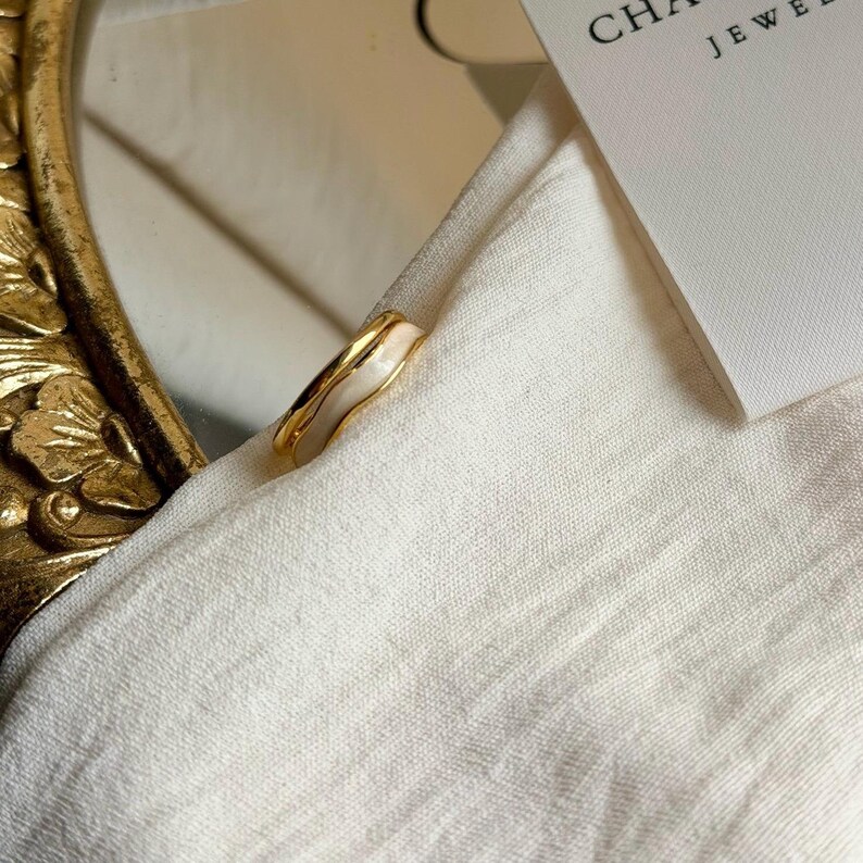 May include: A gold-toned ring with a white enamel inlay. The ring is set against a white fabric background, with a glimpse of a decorative mirror frame and a jewelry tag. The ring's design is organic and modern.