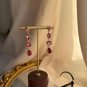 Op de afbeelding: Een paar goudkleurige oorbellen met een verticale opstelling van drie roze en rode gefacetteerde kristallen. Elke oorbel bestaat uit een ronde steen, een rechthoekige steen en een druppelvormige steen die aan een gouden metalen ketting hangt.