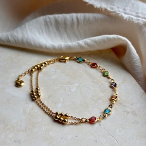 Puede incluir: Una pulsera de doble cadena en tono dorado que presenta una serie de pequeñas cuentas metálicas redondas y una secuencia de piedras preciosas de colores engastadas en tonos rojo, azul, verde y morado, con una cadena fina y un pequeño dije redondo.