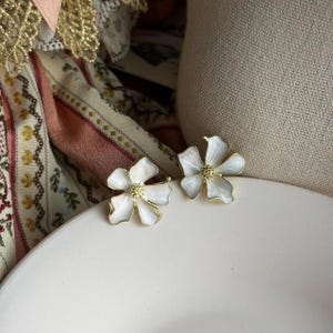 Peut inclure: Une paire de boucles d'oreilles en forme de fleurs à cinq pétales. Chaque boucle présente des pétales en émail blanc avec un contour en métal doré et un centre texturé doré. Le design est symétrique, avec des bords incurvés et lisses.