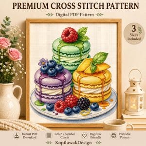 May include: A cross stitch pattern featuring three stacked macarons in green, purple, and yellow on a white plate. The green macaron is topped with a raspberry, the purple with blueberries, and the yellow with mango slices. Text reads PREMIUM CROSS STITCH PATTERN, Digital PDF Pattern, and 3 Sizes Included.