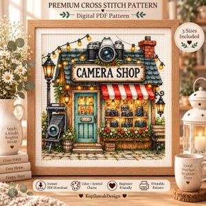 May include: A framed cross stitch pattern featuring a camera shop with a teal door, red and white striped awning, and string lights. The shop displays various cameras in the window and on a stand. Text reads: PREMIUM CROSS STITCH PATTERN.