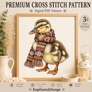 May include: A digital cross stitch pattern featuring a fluffy duckling wearing a knitted scarf with a snowflake pattern in shades of brown, orange, and white. The text reads PREMIUM CROSS STITCH PATTERN, Digital PDF Pattern, 3 Sizes Included, and KopiluwakDesign.