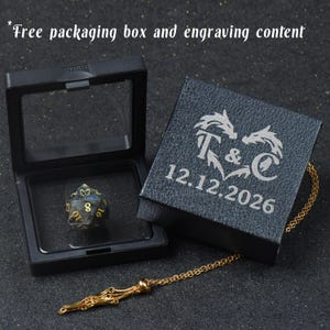 May include: A black display box with a clear lid containing a dark polyhedral die with gold numbers, alongside a matching black box lid engraved with a dragon heart design, the letters T & C, and the date 12.12.2026, plus a gold-toned chain necklace.