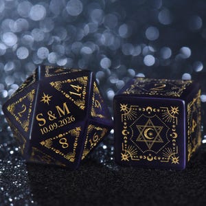 May include: A dark purple twenty-sided die and a six-sided die featuring gold celestial engravings of stars, moons, and geometric patterns. The twenty-sided die displays the letters S & M and the date 10.09.2026 in gold lettering on its surface.