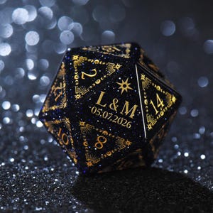 May include: A twenty-sided die with a dark blue glitter finish and gold-coloured engravings. The faces feature celestial motifs including stars and moons, with the custom text L & M and the date 05.07.2026 prominently displayed on one side.