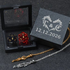 May include: A black display box containing two twenty-sided dice, one dark blue with gold glitter and one translucent red, both featuring the text K&C 12.12.2026. Beside it, a black box engraved with T & C 12.12.2026 and two metal chains in gold and silver.