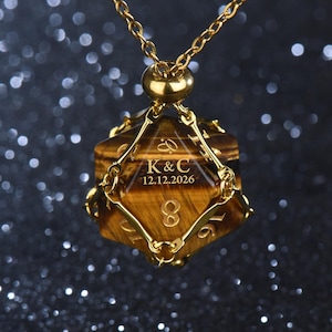 May include: A gold-toned metal cage pendant holding a brown and gold tiger's eye twenty-sided die. The die features the engraved text K & C and the date 12.12.2026. The pendant hangs from a matching gold-toned metal chain link necklace.