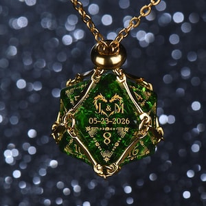 May include: A gold-toned metal cage pendant holding a translucent green twenty-sided die. The die features gold-engraved text reading J & M, the date 05-23-2026, a heart-shaped dragon motif, celestial symbols, and the number 8 on its front face.