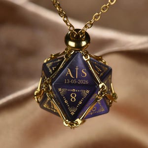 May include: A gold-toned metal cage pendant shaped like a twenty-sided die, suspended from a matching chain. The dark purple die features gold engraved text reading A|S and 13-05-2026, along with the number 8 and celestial star motifs.