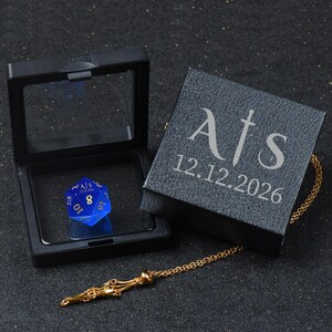 May include: A blue translucent twenty-sided die with gold numbering and the text A|S 12.12.2026, resting in an open black display box. Beside it sits a black box lid engraved with A|S and 12.12.2026, attached to a thin gold-toned metal chain.
