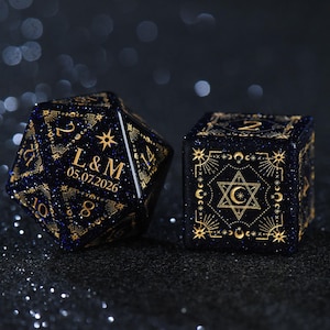 May include: A dark blue glittery twenty-sided die and a six-sided die featuring gold celestial engravings. The twenty-sided die displays the text L & M and the date 05.07.2026. The six-sided die features a central star and crescent moon design.