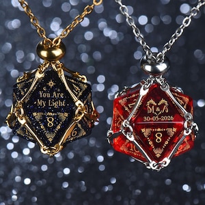 May include: Two polyhedral d20 dice pendants on chains. The left is dark blue with gold glitter, inscribed with 'YOU ARE MY LIGHT' and the number 8. The right is translucent red, inscribed with 'M&A', '30-05-2026', and the number 8 in gold.
