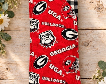Puede incluir: Una funda de tela rectangular con fondo rojo y un patrón repetitivo del logotipo de Georgia Bulldogs, el texto UGA, el texto BULLDOGS, el texto GEORGIA y un gráfico de casco de fútbol americano. El reverso muestra un patrón de cuadros vichy en blanco y negro.