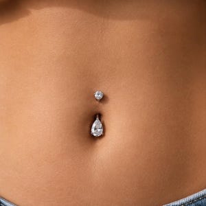 May include: A silver-toned navel piercing ring featuring a small round clear gemstone at the top and a larger pear-shaped clear gemstone dangling at the bottom. The jewelry is positioned within the navel, showcasing a metallic finish and faceted stones.