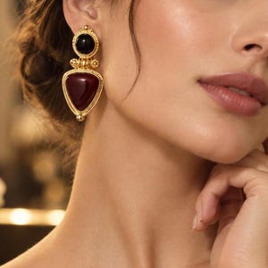 May include: A pair of gold-toned drop earrings featuring a circular black stone at the top and a larger, triangular deep red stone at the bottom. The metal settings have a textured, beaded edge detail, creating a layered, geometric design.