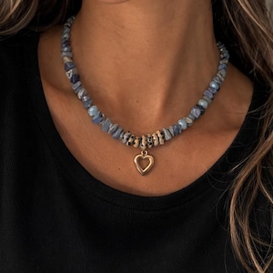 Può includere: Una collana con perline di pietra blu e grigia e un ciondolo a forma di cuore dorato. La collana è indossata su un top nero, creando un contrasto che mette in risalto il gioiello.