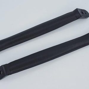 Puede incluir: Dos manijas negras y alargadas de goma, con extremos redondeados y orificios de montaje. Una manija tiene un solo orificio, la otra tiene dos. Las manijas son probablemente para un equipo o mueble.