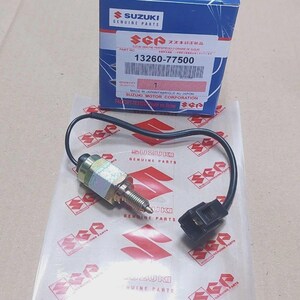 Puede incluir: Un interruptor Suzuki Genuine Parts nuevo con cable y conector negros. El número de pieza 13260-77500 es visible en la caja. La imagen también muestra el logotipo y el texto de Suzuki Genuine Parts.