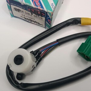 Op de afbeelding: Een nieuwe Patron Kabelschakelaar met een zwarte kabel, een witte en zwarte schakelaar en een groene connector. De doos op de achtergrond heeft de tekst "Patron Cable Switch CS-112037".