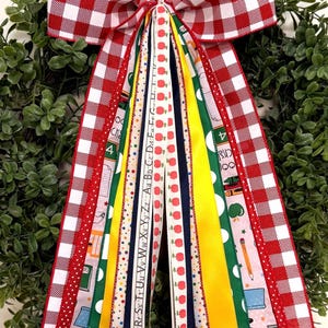 May include: A decorative bow made of multiple ribbons featuring a red and white gingham pattern, solid yellow, solid green, and navy blue strips. Several ribbons display school-themed prints including red apples, pencils, and the alphabet letters.