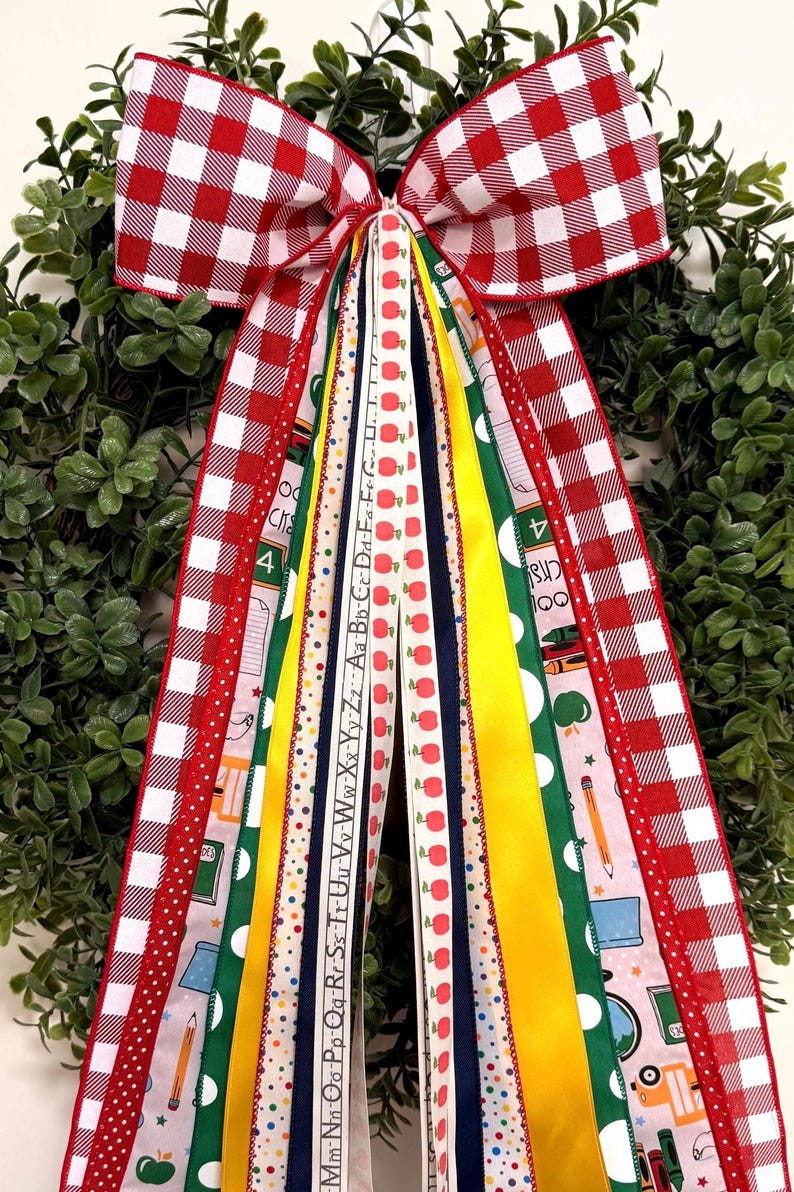 May include: A decorative bow made of multiple ribbons featuring a red and white gingham pattern, solid yellow, solid green, and navy blue strips. Several ribbons display school-themed prints including red apples, pencils, and the alphabet letters.
