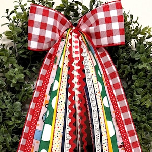 May include: A decorative wreath bow featuring a red and white gingham pattern at the top with long, cascading ribbons in various school-themed designs, including alphabet letters, colorful polka dots, pencils, and apples on green and white fabric.
