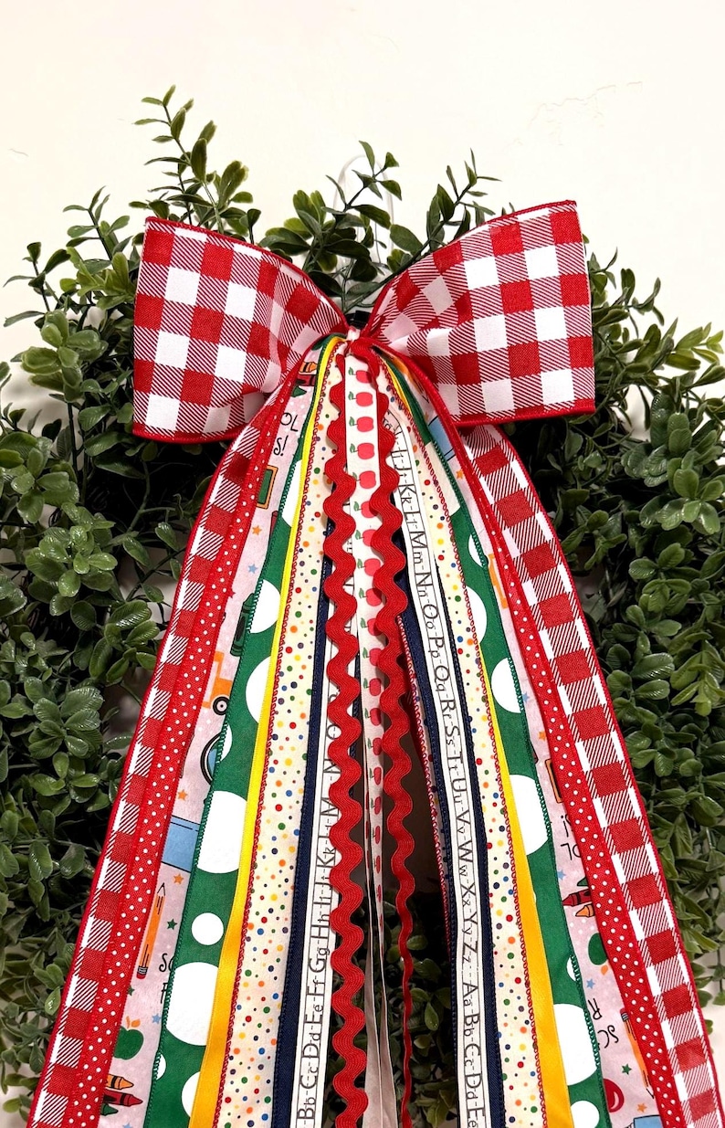 May include: A decorative wreath bow featuring a red and white gingham pattern at the top with long, cascading ribbons in various school-themed designs, including alphabet letters, colorful polka dots, pencils, and apples on green and white fabric.