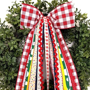 May include: A decorative wreath featuring a large red and white gingham bow with long, cascading ribbons. The ribbons display various patterns including colorful polka dots, solid yellow, navy blue, red rickrack, and school-themed motifs.